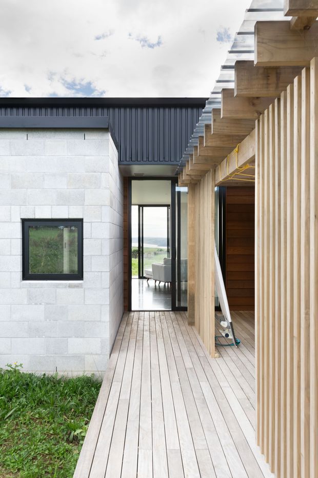 A contemporary covered timber deck or walkway runs alongside a house, with pale timber decking, vertical timber screening, and exposed timber pergola beams. The adjacent wall is constructed from light concrete masonry blocks with a dark metal-clad roof edge. Glazed doors lead into an interior seating area, while open framing beyond reveals a green landscape.