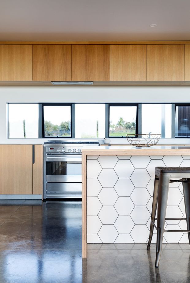 Bright modern kitchen featuring extensive light timber cabinetry, a large island with a white hexagonal tile face, stainless-steel range cooker, dark metal-framed windows, and a polished concrete floor. A metal bar stool and wire fruit bowl add industrial accents.