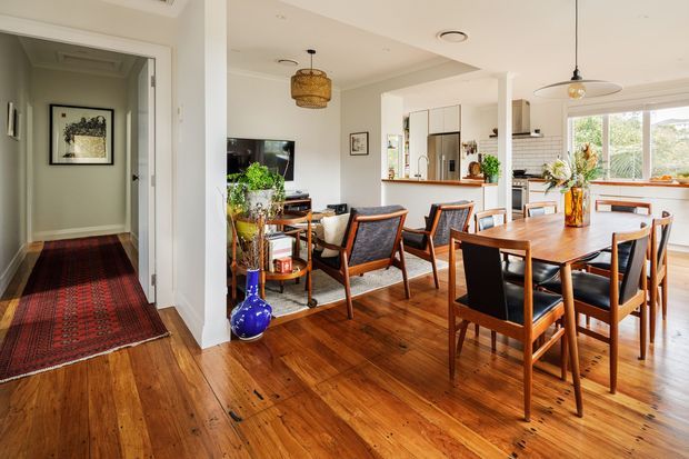 Open-plan dining and living area with a timber dining table and chairs, mid-century-style lounge chairs, polished timber flooring, white walls, and abundant natural light. A kitchen is visible at the rear, while a carpeted hallway extends to the left.