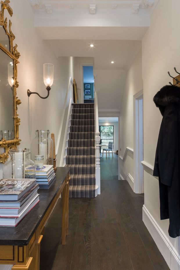 Elegant residential hallway and stairway with dark timber flooring, a striped carpet runner on the stairs, white painted walls and ornate ceiling cornice detailing. A richly carved gilt mirror, console table, glass vessels and stacked books occupy the left side, while a coat rack and dark coat sit on the right. The hall leads toward a brighter room with a glimpse of a dining area beyond.