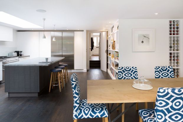 Bright open-plan kitchen and dining area with a large dark island, white cabinetry, stainless-steel appliances, timber dining table, patterned upholstered chairs, dark timber flooring, and extensive built-in shelving and wine storage. A hallway and additional rooms are visible beyond the kitchen.