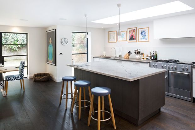 Bright contemporary kitchen with a large marble-look island, dark timber-toned cabinetry, three blue upholstered stools, stainless-steel range cooker, pendant lights, skylight, and framed artwork. A small dining area is visible at the left.