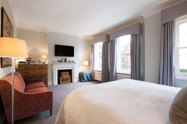 A spacious, traditionally styled bedroom with a large upholstered bed, a fireplace with white mantel, wall-mounted television, dresser, armchair, floor lamp, and tall bay-style windows dressed in grey curtains. The room has soft neutral finishes, carpet flooring, decorative ceiling cornices, and warm layered lighting.