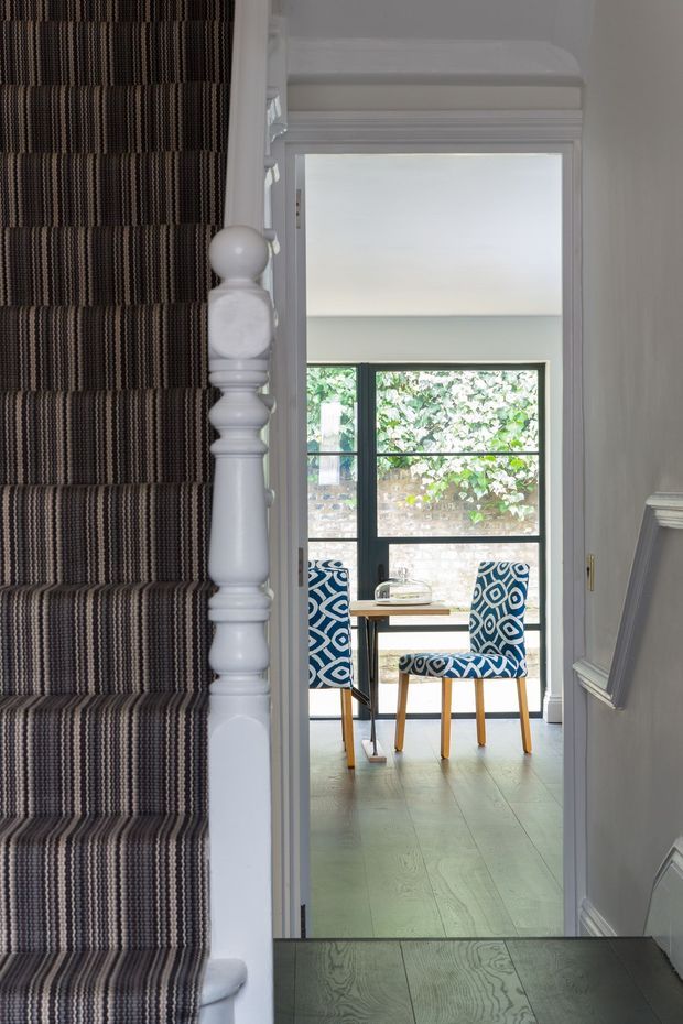 A view from a stairway into a bright dining area. The staircase has a white painted turned balustrade and dark striped carpet runner, while the adjoining room contains a small wooden dining table with blue-and-white patterned upholstered chairs, pale timber flooring, and large black-framed glazing. Traditional wall mouldings and muted grey walls create a refined, layered interior.