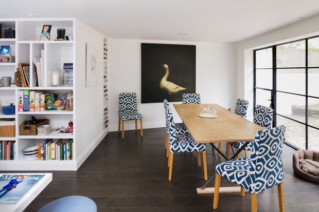 Bright, contemporary dining room with a large timber dining table surrounded by patterned upholstered chairs. White built-in open shelving displays books and objects along the left wall, while a large black-framed glazed door or window provides abundant natural light on the right. Dark timber flooring, white walls, and a large swan artwork create a crisp, graphic interior.
