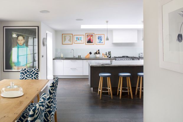 Bright open-plan kitchen and dining area with white flat-panel cabinetry, dark stone benchtops, a large central island with three blue stools, and dark timber flooring. A timber dining table with patterned upholstered chairs occupies the foreground, while colourful framed artwork and a large framed photograph add visual interest. The space is contemporary, clean, and naturally lit.
