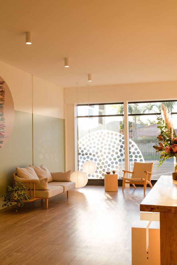 Warm, light-filled contemporary living room with pale peach and muted sage walls, timber flooring, a low upholstered sofa, timber-framed lounge chairs, globe pendant lights, and a substantial timber console or bench. Large windows overlook a garden and feature a circular dotted privacy treatment, while indoor planting and colourful artwork add character.