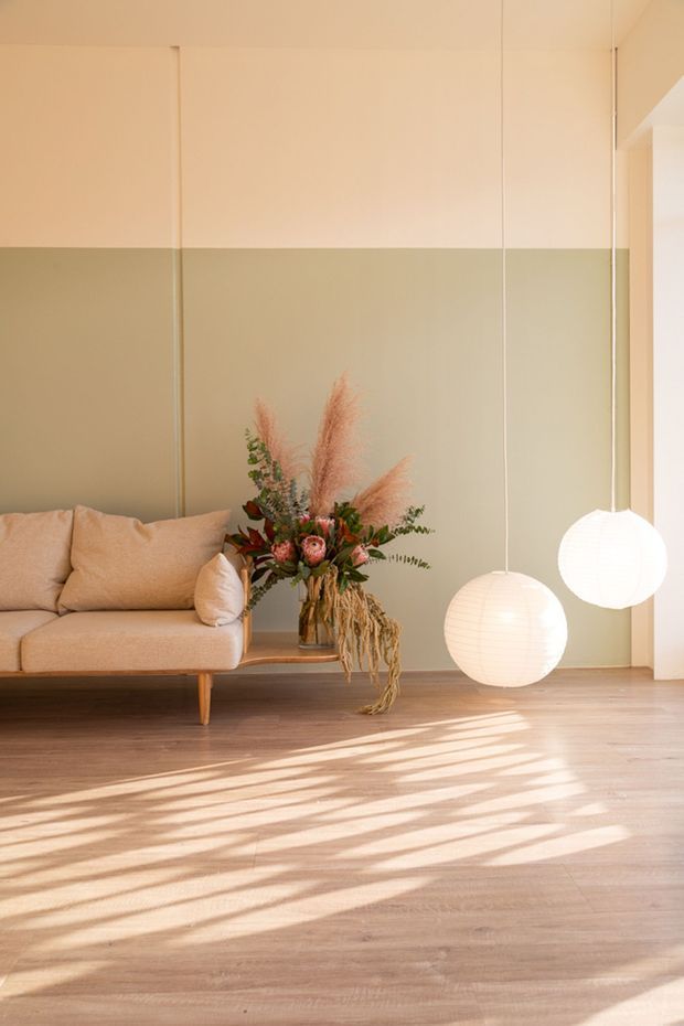 A softly lit minimalist living room with a low beige upholstered sofa, pale sage-green and warm white two-tone walls, natural timber flooring, and two suspended round white pendant lights. A vase of dried and fresh botanical stems sits beside the sofa, while strong sunlight casts patterned shadows across the floor.
