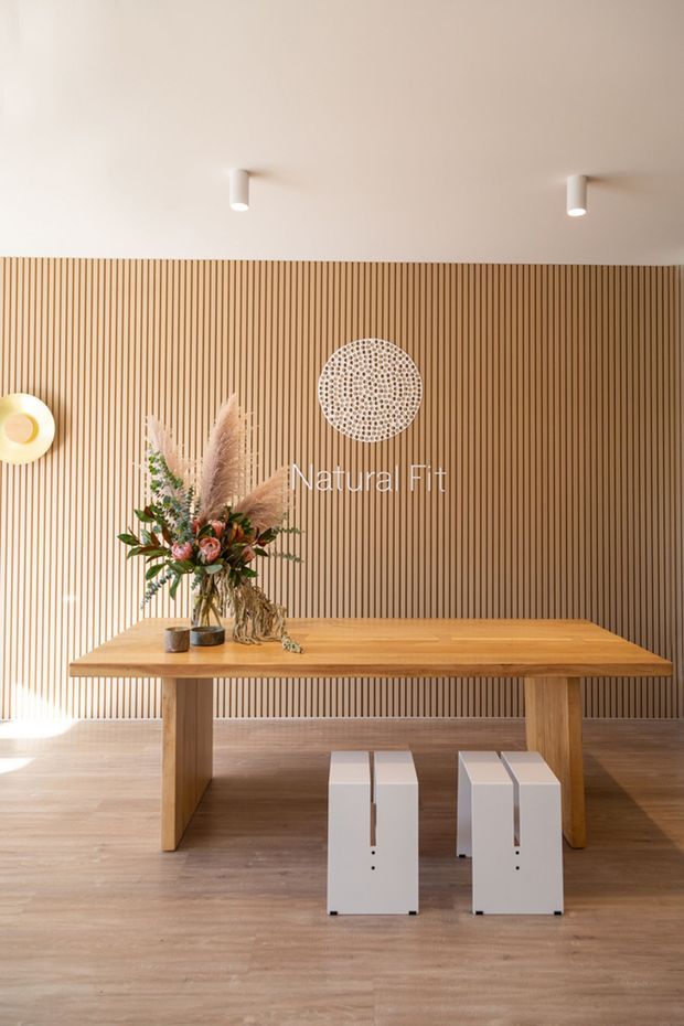 Warm, minimalist interior featuring a long solid timber table with trestle-style legs, paired white block stools, and a floral arrangement. The main feature is a full-height vertical timber slat wall with a circular decorative wall piece, set against a white ceiling with simple recessed-style cylindrical spotlights. Timber flooring and abundant warm natural light create an inviting, natural atmosphere.