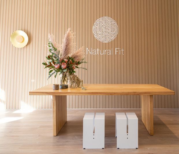 A warm, minimalist entryway or hallway vignette featuring a solid timber console table against a full-height vertical timber batten wall. Two small white stools sit beneath the table, while a floral arrangement, sculptural wall art, and a round wall light add decorative accents. Natural light falls across the timber floor and wall.