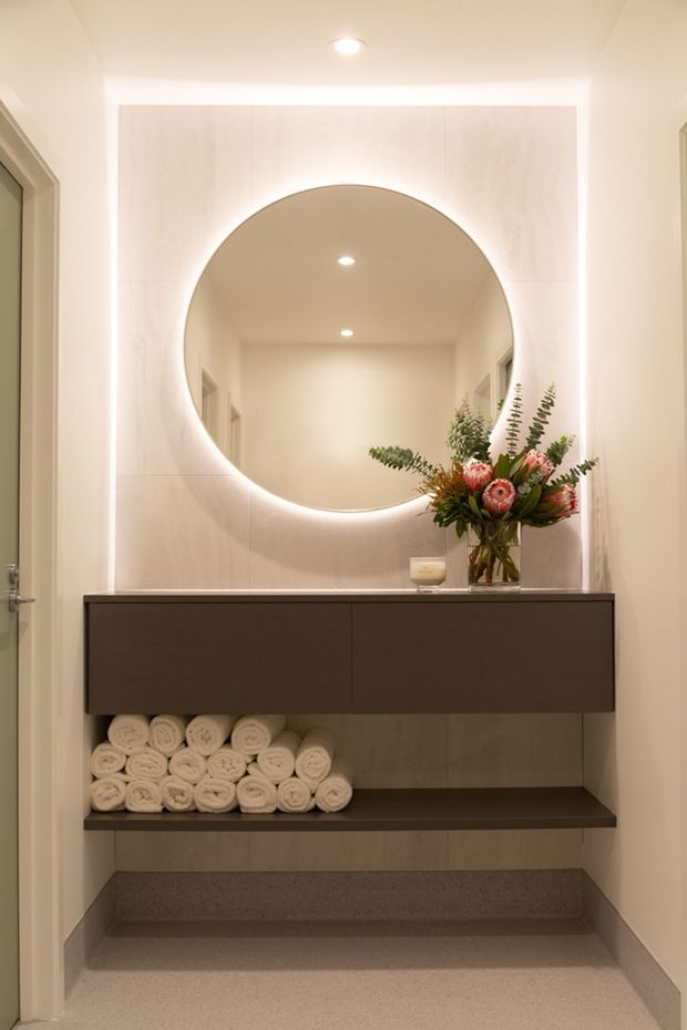 A refined bathroom vanity area with a floating dark wood-toned cabinet, open lower shelf, integrated warm LED lighting, and a large round backlit mirror set into pale stone-look wall panels. Rolled white towels and a vase of flowers sit on the vanity, with a softly lit hallway reflected in the mirror.