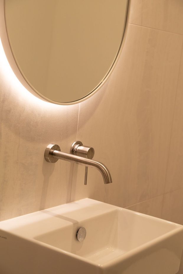 Close-up of a refined bathroom vanity featuring a wall-mounted brushed metal tap over a white rectangular basin. The basin sits against warm beige stone or tile wall surfaces, with a large rounded-oval mirror above and soft concealed lighting creating a warm glow.