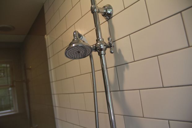 Close-up of a traditional chrome shower fitting with a fixed showerhead, handheld shower hose, and exposed riser pipe mounted against a wall of white rectangular subway tiles with dark grout. A dimly lit bathroom area and window are visible in the background.