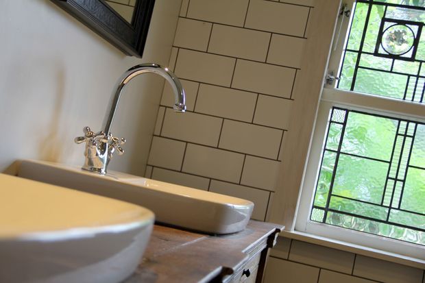 Close-up of a traditional bathroom vanity featuring a white ceramic vessel basin in the foreground, a second white basin with an ornate chrome tap, a timber vanity top, cream rectangular subway wall tiles with dark grout, and a decorative leaded-glass window. The image has a warm, heritage-inspired character.