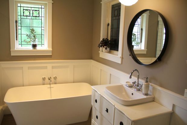 A traditional bathroom with a freestanding white bathtub beneath a decorative leaded-glass window, white-painted wainscoting, and a white vanity with an integrated basin and polished metal fixtures. A large round black-framed mirror and a narrow decorative interior window add contrast and character. The room has warm taupe walls and soft, natural lighting.