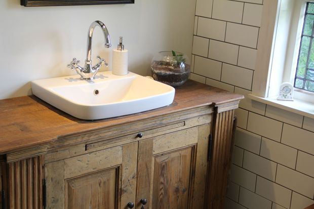 A warmly styled bathroom featuring a freestanding white vessel basin on a rustic timber vanity with paneled cabinet doors, fluted side details, and a wood countertop. The walls are finished in white rectangular subway tiles with dark grout, and a chrome gooseneck tap, soap dispenser, framed mirror edge, and window complete the space.