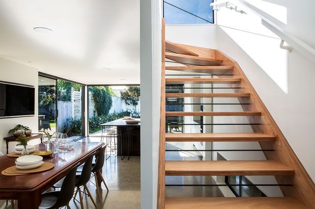 Bright contemporary open-plan interior combining a dining area with a modern stairway. The dining space has a dark timber table, upholstered dining chairs, pendant-free recessed lighting, and large glazed doors opening to a landscaped courtyard. Adjacent is an open-riser staircase with warm timber treads, slim dark metal supports, and a white balustrade, illuminated by strong natural light from high-level glazing.