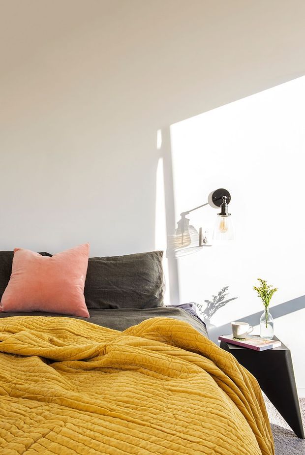 Bright, minimal bedroom vignette featuring a low bed with charcoal grey bedding and a mustard-yellow quilt, accented by a coral-pink pillow. A black wall-mounted swing-arm light, small black bedside table, white cup, and vase with greenery sit against a plain warm-white wall. Strong sunlight creates crisp shadows and a fresh, inviting atmosphere.