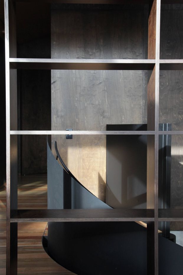 A close architectural interior view through a dark timber and plywood open-screen shelving or partition. Behind it is a sculptural black curved staircase with a compact landing and dark wood flooring, creating a layered, moody composition with strong natural light and shadow.