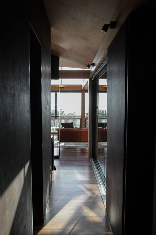 A narrow, dark contemporary hallway leads toward a bright living area and glazed balcony or terrace. The corridor has dark timber or stained wall surfaces, a smooth concrete ceiling with compact spotlights, polished timber flooring, and extensive black-framed glazing. A rust-coloured sofa and exposed timber structure are visible beyond, with strong sunlight creating linear shadows across the floor.