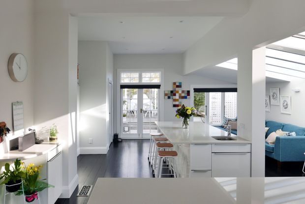 Bright open-plan contemporary interior viewed from the kitchen, with a large white island and waterfall-style benchtop, integrated cabinetry, bar stools, dark timber flooring, and a living room visible beyond. Strong daylight enters through glazed doors and a roof skylight; white walls and ceilings create an airy, clean atmosphere, with a vivid blue sofa and colourful wall art as accents.