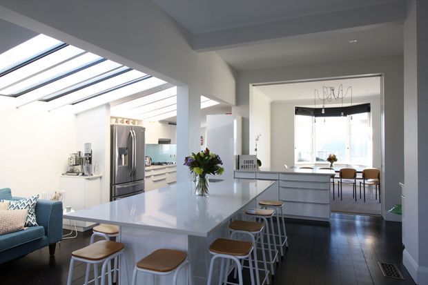 Bright open-plan kitchen and dining area with a large white island, integrated seating, white cabinetry, stainless-steel refrigerator, skylights, and a dining table beyond. A glimpse of a blue upholstered living area is visible at left. The space has dark timber flooring, white walls, abundant natural light, and a clean contemporary fit-out.