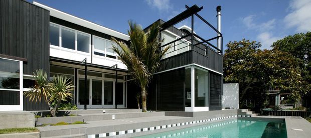 A contemporary multi-level residential exterior viewed from the pool area, featuring dark vertical cladding, extensive white-framed glazing, a projecting upper-level structure with an exposed black steel pergola, and a tiled swimming pool in the foreground. Tropical planting and mature trees surround the outdoor setting.