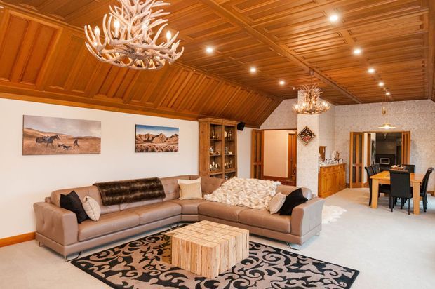 Warm, richly detailed open-plan living and dining area with a large sectional sofa, patterned rug, timber coffee table, built-in display cabinetry, and a dining table beyond. The room features extensive dark honey-toned timber wall and coffered ceiling panelling, angled timber ceiling sections, ornate antler-style chandeliers, and light carpet flooring. Artwork and decorative furnishings contribute to a lodge-inspired, traditional atmosphere.