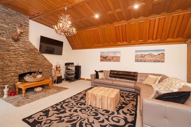 Warm, richly detailed living room with a coffered timber ceiling and matching timber wall trim, a stacked-stone fireplace wall, leather sectional seating, patterned rug, rustic timber coffee table, and sculptural chandelier. Neutral furnishings and desert landscape artwork complement the strong natural materials.