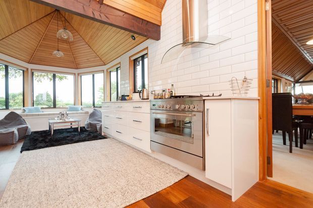 A bright open-plan kitchen and living area with white subway-tile walls, white cabinetry, a stainless-steel range cooker and glass rangehood. Warm timber flooring, a timber-lined vaulted ceiling with exposed beams, and extensive corner glazing create a light, natural interior. A dining area is visible through the timber-framed opening on the right.