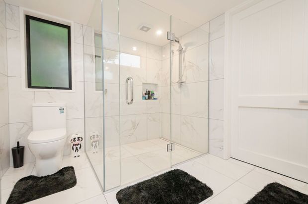 Bright, contemporary bathroom featuring a fully tiled walk-in shower enclosed by clear glass, white marble-look wall and floor tiles, a wall-hung toilet, recessed shower shelving, chrome fixtures, and a frosted window. Dark textured bath mats provide contrast against the predominantly white finishes.