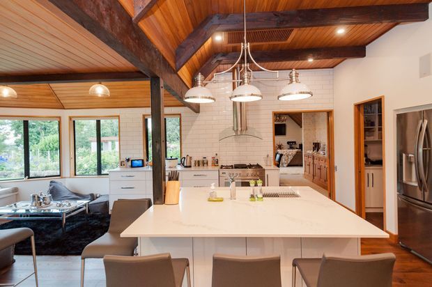 Open-plan kitchen with a large white island and integrated seating, white subway-tile walls, stainless-steel appliances, timber cabinetry, and a dramatic exposed timber-beam ceiling. A dining and living area is visible to the left, with extensive glazing and warm natural light.