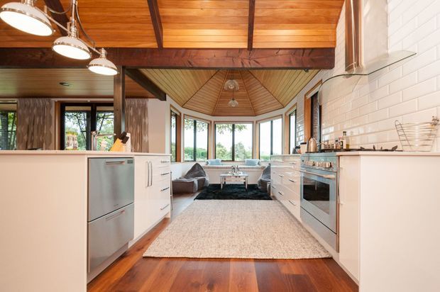 Open-plan kitchen leading into a living area, featuring white cabinetry, stainless-steel appliances, a white brick-tile splashback, rich timber flooring, and a warm timber-lined vaulted ceiling with exposed dark beams. Large windows provide abundant natural light and views of greenery.