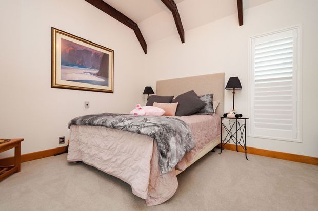 A cozy bedroom with a double bed dressed in pale pink bedding and a grey patterned throw, dark accent pillows, matching bedside lamps, framed artwork, beige carpet, white walls, plantation shutters, and exposed dark timber ceiling beams.