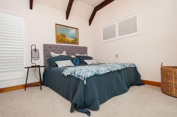 A bright, comfortably furnished bedroom with a centered bed dressed in dark teal bedding, patterned cushions, and a textured blue throw. The room has a tufted grey upholstered headboard, pale walls, beige carpet, white plantation shutters, dark exposed ceiling beams, timber skirting, and a wicker basket. A small black side table and decorative lamp sit beside the bed, with artwork above the headboard.