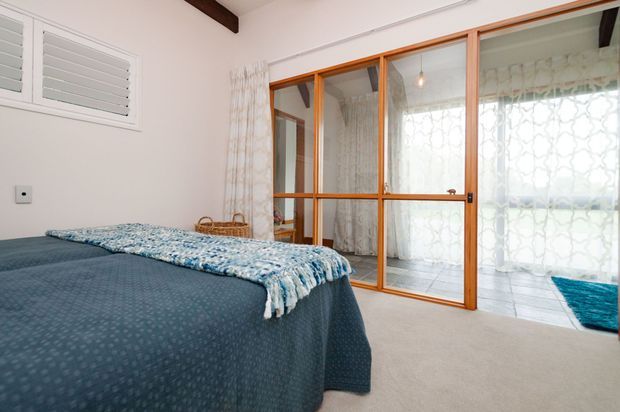Bright bedroom with a neatly made bed, pale carpet, white walls, timber ceiling beam, louvred window shutters, and a timber-framed glass sliding partition opening to a light-filled adjoining space. Sheer patterned curtains and blue textiles add a restrained coastal feel.