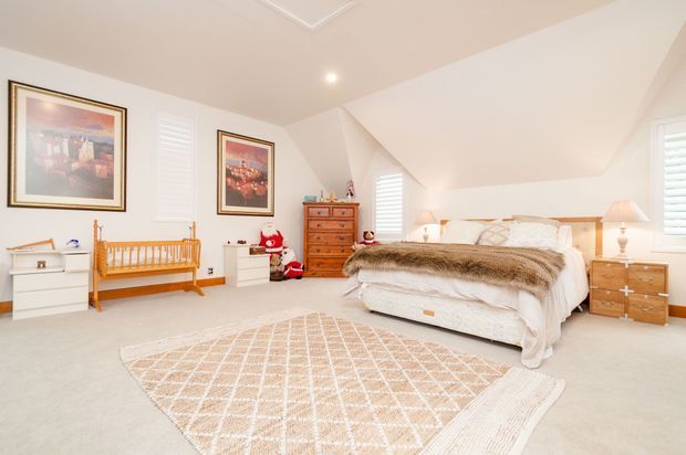 Bright, spacious attic-style bedroom with a sloped ceiling and dormer-like angled walls. The room contains a large upholstered bed with layered white bedding and a fur throw, matching bedside tables and lamps, a timber chest of drawers, a timber crib, children's toys, framed artwork, white plantation shutters, and a large neutral patterned rug over pale carpet.