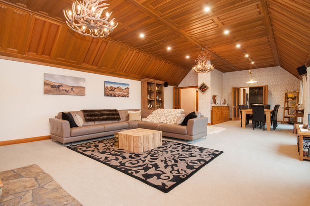 A spacious open-plan living and dining area with a dramatic warm timber coffered and pitched ceiling, recessed lighting, and antler-style chandeliers. The room has neutral carpet, a large patterned rug, two sofas arranged around a rustic timber coffee table, built-in timber shelving, a dining table, and stone flooring at the foreground. White textured walls and timber detailing create a rustic lodge-inspired interior.