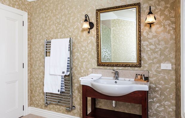 Traditional bathroom with ornate beige floral wallpaper, a white pedestal-style basin set into dark timber joinery, chrome heated towel rail, decorative framed mirror, wall sconces, and a white paneled door.