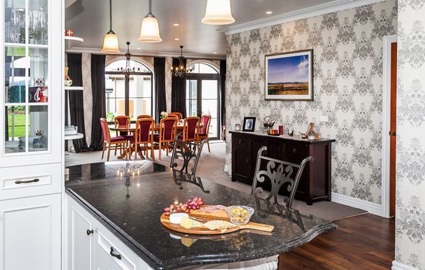A refined open-plan interior viewed from a kitchen island, with dark polished stone benchtops and white cabinetry in the foreground. The space opens into a formal dining area with a large wooden table and red upholstered chairs, tall arched windows and doors with dark curtains, ornate pendant and chandelier lighting, patterned grey wallpaper, dark timber flooring, and a traditional sideboard. A framed landscape artwork and decorative accessories complete the room.