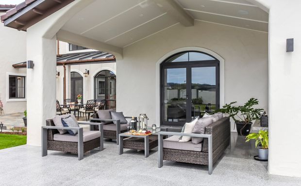 Covered outdoor living terrace with a Mediterranean-inspired stucco facade, arched openings, dark-framed arched glazing, tiled roof edges, and a coordinated wicker seating arrangement around a low coffee table. The space opens toward a landscaped courtyard and appears designed as a sheltered outdoor lounge.