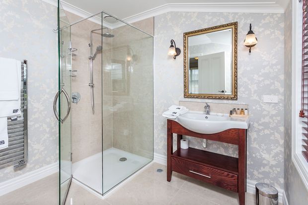 A bright traditional bathroom with a frameless glass corner shower, white ceramic basin on a dark timber vanity, pale patterned wallpaper, marble-look flooring, ornate framed mirror, wall sconces, and a heated towel rail.