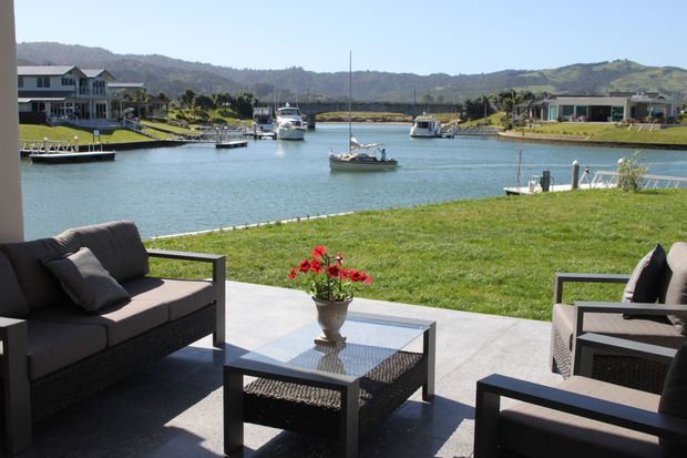 A waterfront outdoor patio or terrace overlooking a calm marina canal, with dark contemporary outdoor lounge seating, a low coffee table, and a potted flowering plant. The landscaped lawn slopes gently toward the water, with boats, neighbouring waterfront homes, and green hills beyond.
