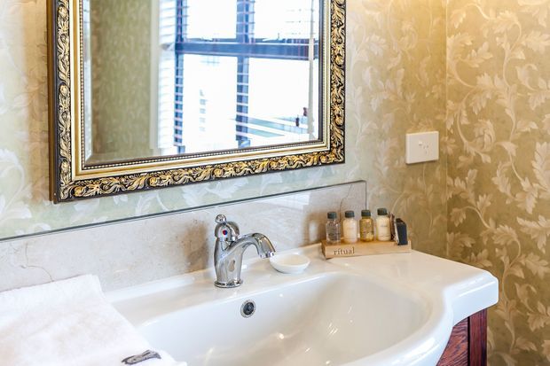 Close-up view of a traditional bathroom vanity with a white ceramic basin, chrome tapware, ornate framed mirror, marble-look backsplash, and beige floral wallpaper. Toiletries sit on a small tray beside the basin, and a towel is visible in the foreground.