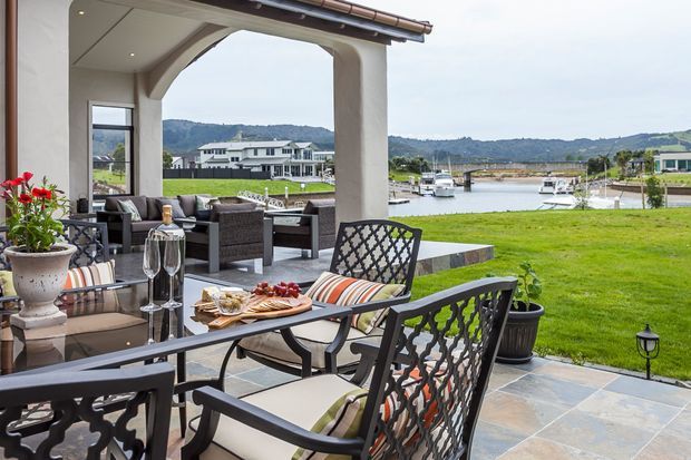 A covered outdoor terrace with a formal dining table and ornate metal chairs, adjacent to a lounge setting and opening onto a manicured lawn and waterfront marina. The terrace has pale stone-look floor tiles, stucco arches, dark-framed glazing, and a tiled roof edge. The scene feels refined and inviting, with boats and neighbouring homes visible across the water.