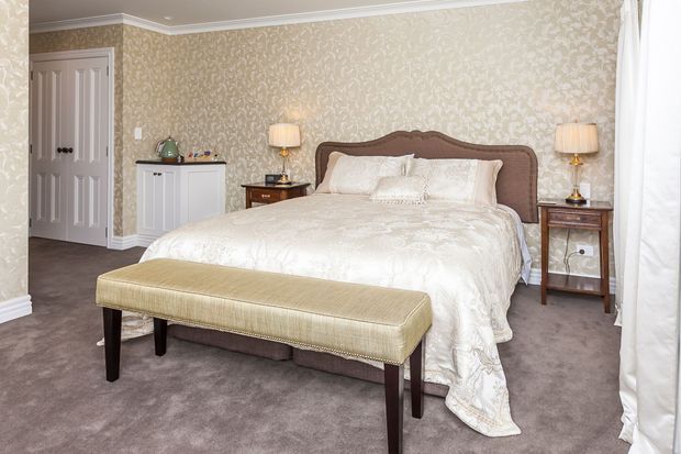 A traditional-style bedroom with a large upholstered bed, cream patterned bedding, matching bedside tables and lamps, a cushioned bench at the foot of the bed, taupe carpeting, ornate beige patterned wallpaper, white crown moulding, and a white built-in cabinet near the doorway. The room is softly lit and coordinated in warm neutral tones.