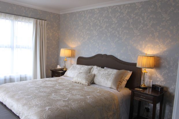 A refined bedroom with a large dark wood bed featuring a curved, carved headboard, dressed in a pale patterned duvet and layered decorative pillows. The walls are covered in ornate grey floral wallpaper, complemented by classic white crown moulding, sheer curtains, and matching traditional bedside tables with warm table lamps.