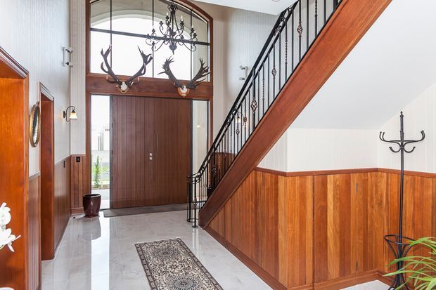 A bright, elegant residential entry foyer with a tall timber front door beneath an arched transom window, polished light stone flooring, dark timber wall panelling, and a prominent timber staircase with ornate black wrought-iron balustrades. Antler wall decor and a traditional chandelier reinforce the formal, classic character.