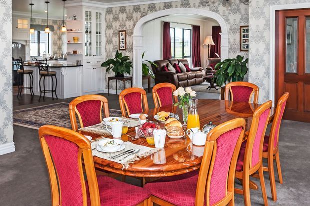 A richly detailed traditional dining room with a large polished timber dining table surrounded by upholstered red dining chairs. The room opens through an arched opening to a formal living room, while a white kitchen with pendant lighting and glass-front cabinetry is visible to the left. Patterned wallpaper, dark timber flooring, ornate furnishings, and layered textiles create a classic, formal interior.