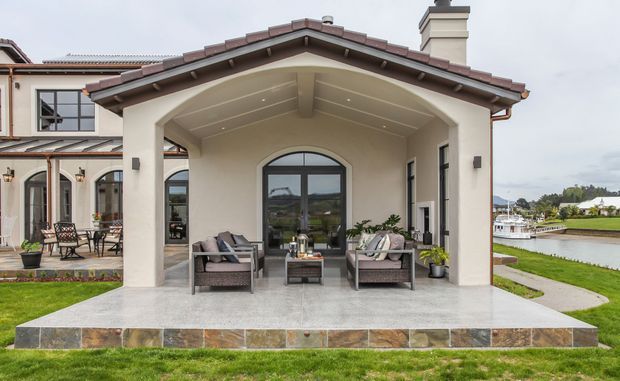 Exterior view of a Mediterranean-inspired home with a covered outdoor living area facing a landscaped waterfront. The sheltered terrace has a vaulted, beamed ceiling, stucco columns, dark-framed glazing, tiled roof edges, stone-clad steps, and outdoor lounge seating.